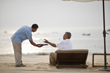 Waiter serving wine to man sitting on sun lounger at beach