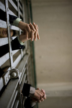 Hands of a young adult businessman and a colleague through bars of a prison cell in a derelict building.