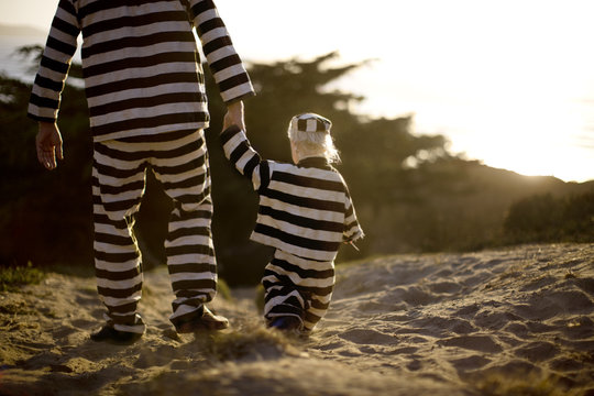Young boy walking hand in hand with his father while wearing a striped prisoner uniform on a beach at sunset.