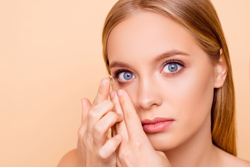 Portrait of pretty, charming, nude, natural girl putting crystalline lens in right eye with forefinger looking at camera isolated on beige background