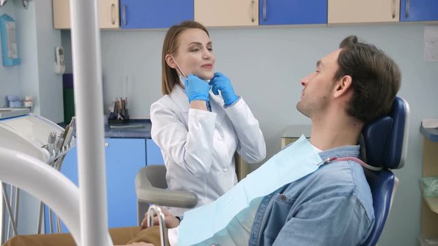 Pretty smiling female dentist in white lab coat and examination gloves putting on dental face mask, taking dental tools and starting check-up on young male patient in dental clinic.