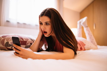 Young brunette in sleepwear can't believe in what she see on her mobile phone while lying on bed at home and