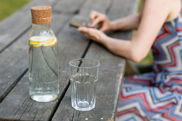 A bottle of water with lemon and a glass. A table and a person with a cell in their hands.