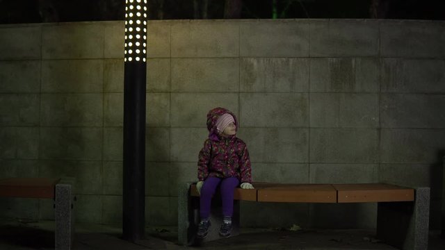 A little girl is sitting on a bench under the light of a lantern at night in a park. The child is lost, worried and waiting for the parents.