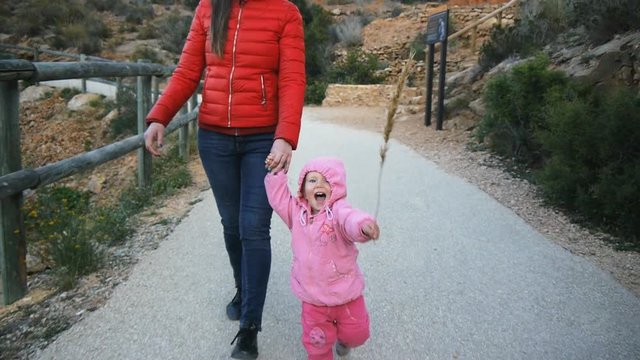 Cute caucasian baby in the hood with herb strow laughing runs holding mom's hand trying to chase the camera in natural park. Happy childhood concept