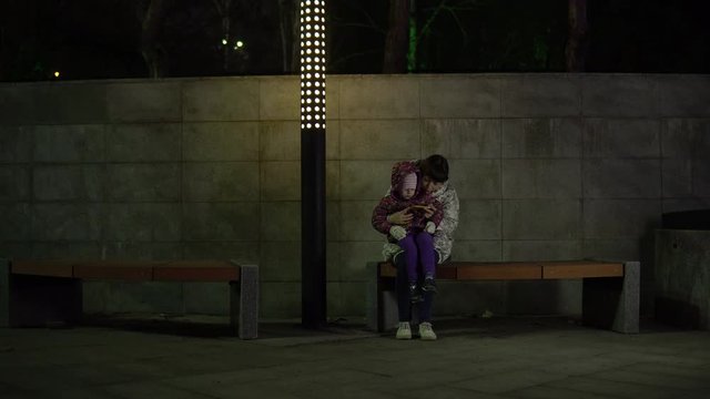 Mother and daughter sit on a bench under the light of a lantern and use the phone at night in the park. A woman is reading to a child.