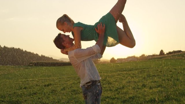 SLOW MOTION, CLOSE UP, LENS FLARE: Male dancer lifts blonde woman in the air and gives her a romantic kiss in the rain. Stunning couple dances passionately in breathtaking rural landscape at sunrise.