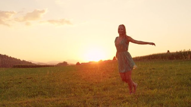 LENS FLARE, COPY SPACE: Happy young dancer spinning outdoors on a pleasant rainy evening. Picturesque summer nature and golden sunrise surround carefree blonde haired woman dancing alone in the rain.