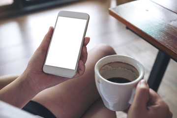 Mockup image of woman holding white mobile phone with blank screen while drinking coffee in cafe