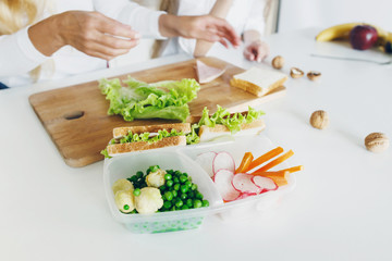 School lunch boxes with sandwich and fresh vegetables on the home kitchen table. In the background, the mother and her daughter are preparing food. Healthy eating concept
