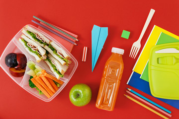 Healthy lunch boxes with sandwich and fresh vegetables, bottle of juice and school supplies on red background. top view. Healthy food concept. Flat lay composition