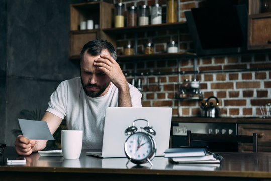 depressed young man looking at photo card in hand while sitting at kitchen with laptop and alarm clock