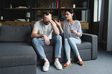 sad young man sitting on couch and looking away while his wife shouting at him and holding credit card