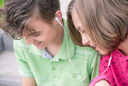 Happy teenage boy and girl with headphones are using gadget, talking and smiling while sitting on the stairs outdoors. Young sister and brother teens playing on mobile phone and listening to music.