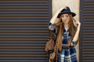 Red hair girl in plaid shirt and vintage hat, with brown backpack pose on the street. Fashion and stylish concept.