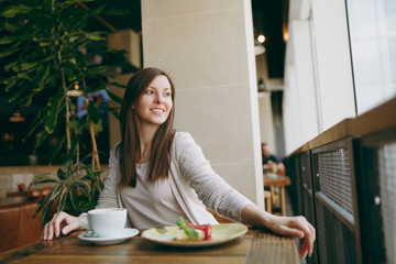 Attractiveyoung woman sitting alone near big window in coffee shop at table with cup of cappuccino, cake, relaxing in restaurant during free time. Young female having rest in cafe. Lifestyle concept.