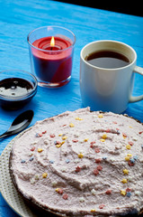 Appetizing pie with a large layer of pink cream. Dessert decorated with caramel stars. A cup of tea, jam and a candle for a romantic tea party. Blue wooden table and black teaspoon.