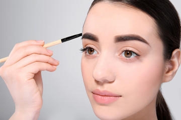 Beautiful woman with perfect eyebrows applying makeup on light background