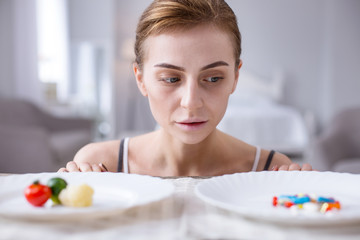 Obraz na płótnie Canvas What to choose. Depressed young woman looking at the pills while standing behind two plates