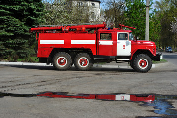 Red fire truck rides along the road from the garage to emergency call, side view with reflection in puddles, against a background of a spring city landscape