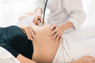 Female Obstetrician doctor with stethoscope listening to pregnant woman baby heartbeat at hospital.