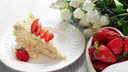 A piece of Napoleon cake and a heart-shaped bowl with a red strawberry, a bouquet of flowers next to it