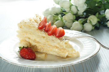 A piece of Napoleon cake on a plate adorned with a red ripe strawberry, a bouquet next to it