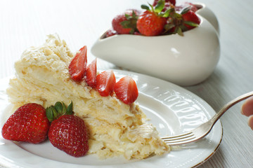 A fork with a piece of cake Napoleon on a plate, decorated with a red ripe strawberry, behind a bowl with berries
