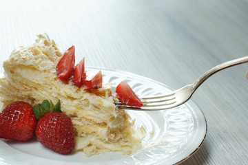 A fork with a piece of cake Napoleon on a plate, decorated with a red ripe strawberry on a table
