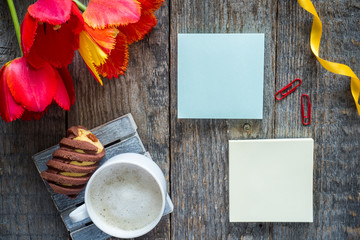 Three colorful tulips on a wooden table. Notepad blank for text. Cup of coffee, cookies