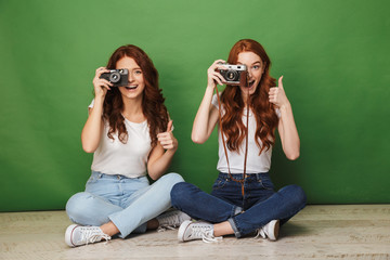 Photo of two redhead women 20s sitting on floor with legs crossed and photopraphing you on retro camera, isolated over green background