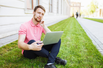 Handsome man sitting on the grass in the city with a laptop and talking on the phone, job search