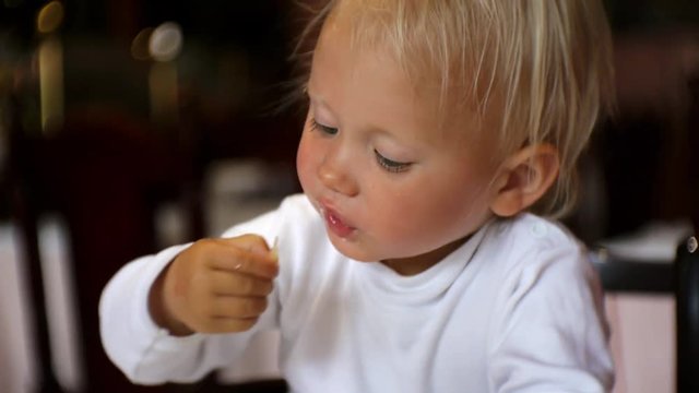 Baby eating food in restaurant. Blonde blue-eyed child in highchair taking piece of food by hand from a plate putting it in the mouth