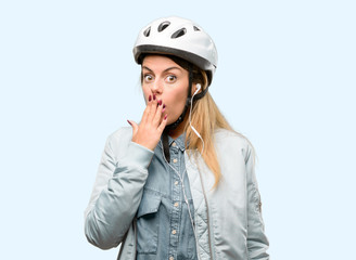 Young woman with bike helmet and earphones covers mouth in shock, looks shy, expressing silence and mistake concepts, scared