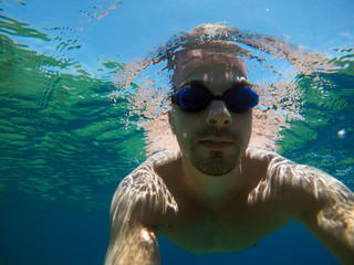 Underwater view of a young diver man swimming in the turquoise sea under the surface with snorkeling mask for summer vacation while taking a selfie with a stick.