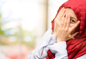 Young arab woman wearing hijab smiling having shy look peeking through her fingers, covering face with hands looking confusedly broadly
