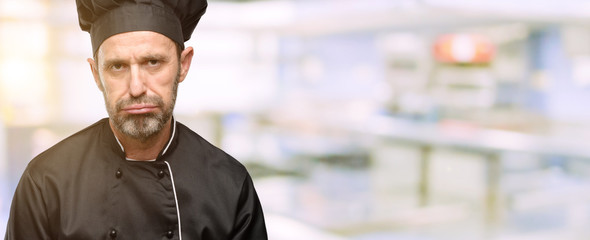 Senior cook man, wearing chef hat with sleepy expression, being overworked and tired at restaurant...