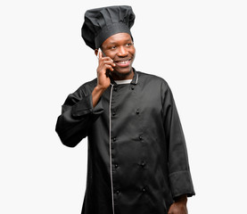 Young black cook wearing chef hat happy talking using a smartphone mobile phone