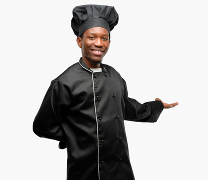 Young black cook wearing chef hat confident and happy with a big natural smile inviting to enter