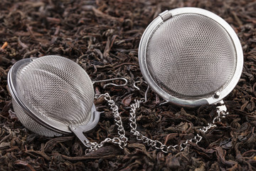 Two tea filter on a chain with black tea on a pile of dry tea leaves.