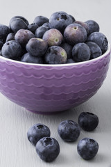 Closeup of ripe blueberries in small purple bowl, five blueberries on the side, against wooden background, healthy eating concept