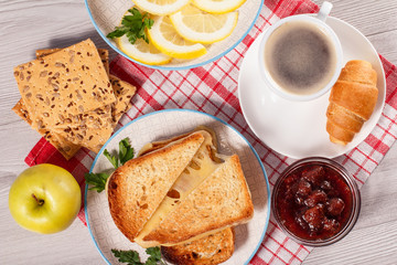 Toasted slices of bread with cheese, apple, cookies with whole grains of sunflower seeds, cut lemon on plate, pup of coffee and croissant on saucer and glass bowl with strawberry jam