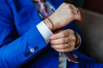 The man sits in an armchair and fixes the cuffs on a white shirt and a stylish blue jacket with expensive gold accessories, watches, rings and rings. The businessman is preparing to meet.