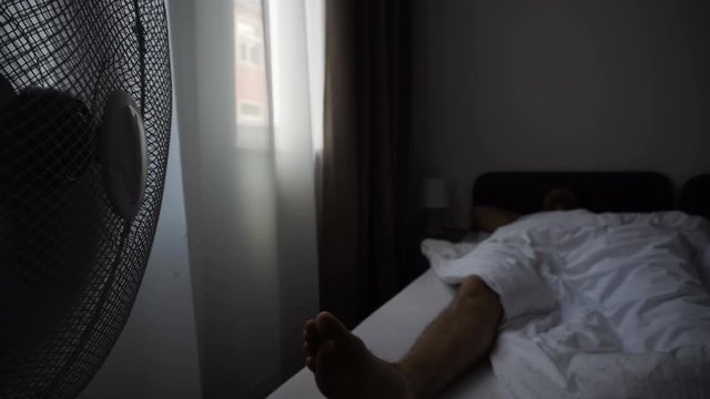 Electric Fan Blowing Air On Man Sleeping In Hotel Room Bed
