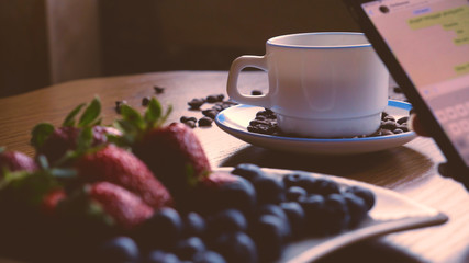 A girl or a woman, writes a message on the phone, a snack, a coffee mug on the table, coffee beans, strawberries and blueberries.