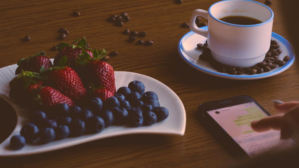 A girl or a woman, writes a message on the phone, a snack, a coffee mug on the table, coffee beans, strawberries and blueberries.