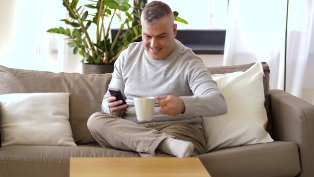 technology, people, lifestyle and communication concept - man with smartphone sitting on sofa at home and drinking coffee