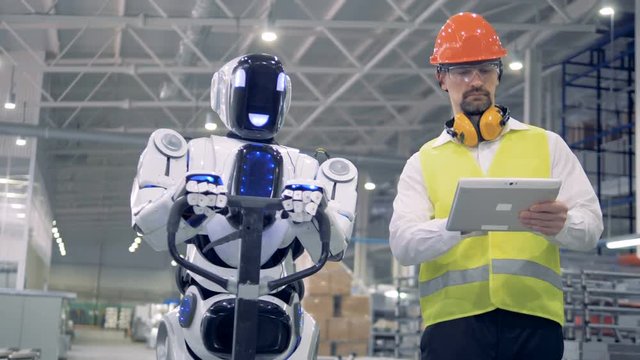 Cyborg is moving a transporting tool along the factory unit while being regulated by a male worker