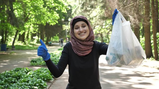 Plogging concept, young girl in hijab runs through park and cleans up garbage, shiwing like sign. 50 fps
