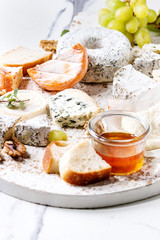 Cheese plate assortment of french cheese served with honey, walnuts, bread and grapes on white wooden serving board over white marble texture background. Close up